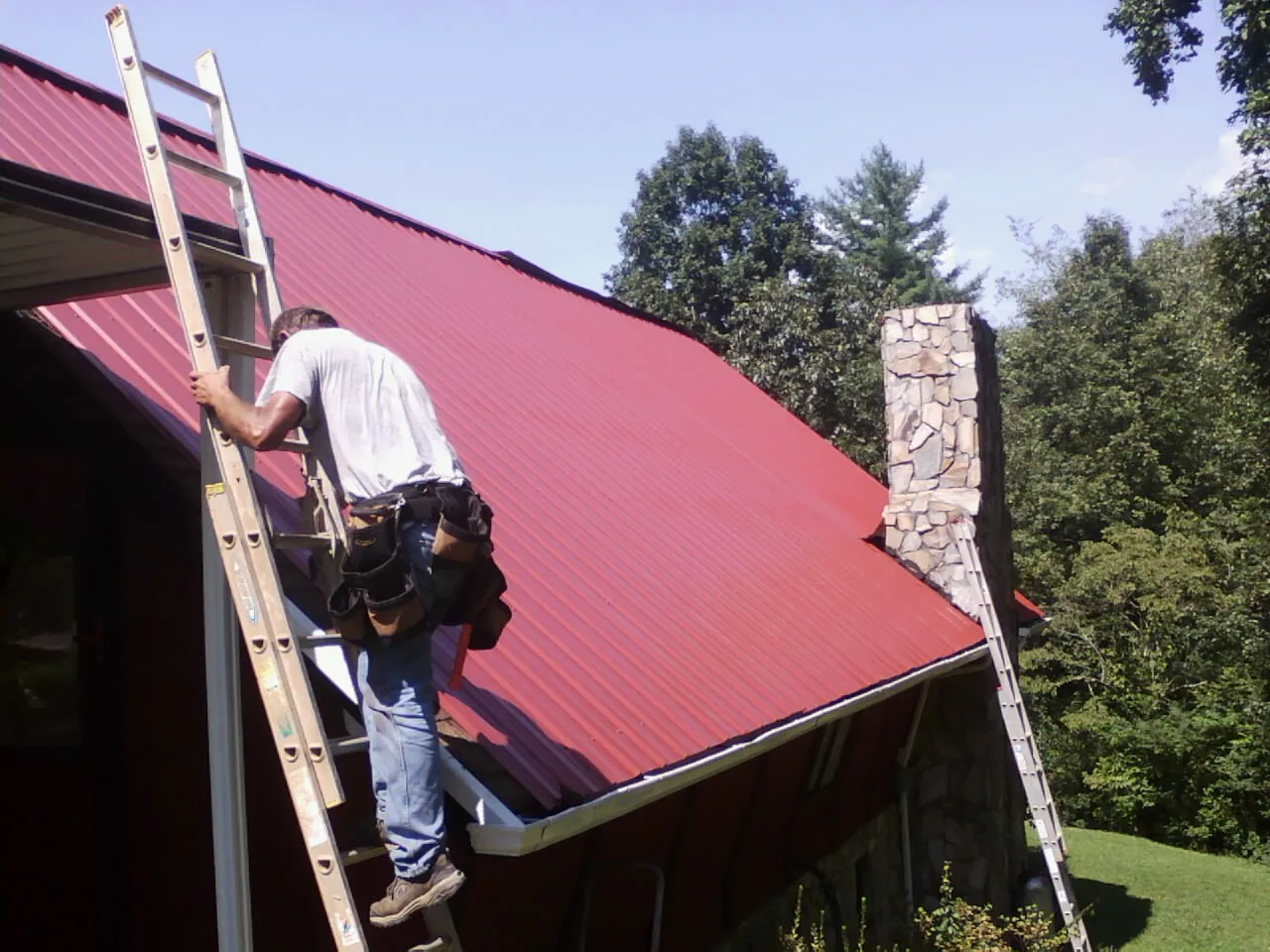 Roofer on a ladder beside a red metal roof and stone chimney — Soffit & Fascia Repair by Roofing Brothers in Seymour